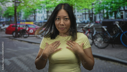 Obraz na płótnie Woman touching chest with both hands on a busy city street near parked bicycles and a red car, looking startled and questioning; surprise self doubt