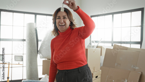 Fotografi Woman holding keys aloft with raised hand and smiling among packed moving boxes in a building; new home joy