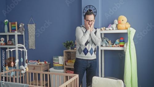 Schilderij op canvas Man with hands clasped near face by wooden crib and hanging mobile in building nursery among shelves and toys; fatherhood bonding tenderness
