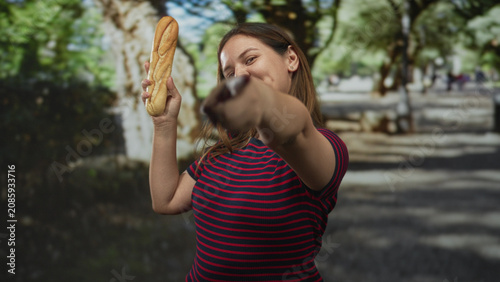 Bild auf Leinwand Woman holding baguette aloft with raised hand and smiling beside benches on a tree lined street under dappled sunlight; playful confidence