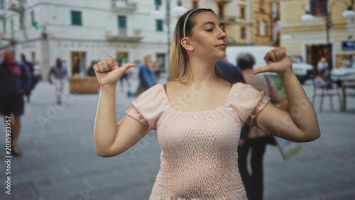 Bild auf Leinwand Woman showing thumbs pointing at herself while wearing a pink smocked dress on a busy street with blurred pedestrians and buildings; confidence assertiveness
