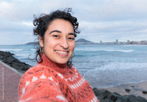 A woman with Arab features and curly hair smiles as she takes a selfie by the sea