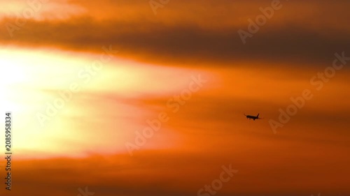 Commercial jet airliner flying against dramatic orange sunset sky. Silhouette of passenger airplane descending in bright golden hour clouds. Telephoto static shot.