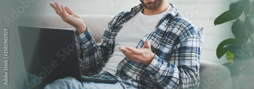 Obraz na plátně Focused Young Man Engaging In Conversation While Using Laptop At Home Comfortably Sitting On Couch