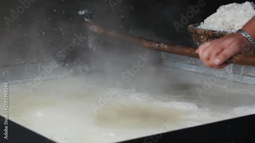 Detail shot of a worker using a wooden tool to stir boiling brine in a large pan during traditional salt production. Thick steam rises as water evaporates to extract natural salt crystals.
