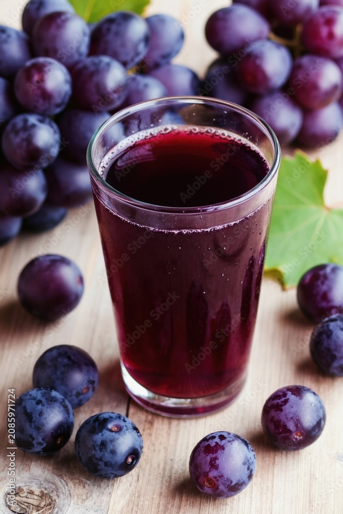 Obraz premium Glass of dark grape juice sits on wooden table with ripe grapes. Freshly squeezed beverage made from whole fruits, healthy drink for refreshment and wellness.