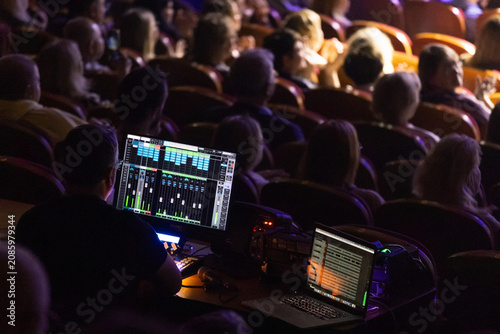 Sound engineer operates mixing console and laptop from backstage during live performance while audience sits in dimly lit auditorium; scene shows illuminated audio control screens, rows of seated