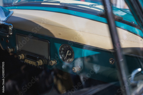 Interior view of vintage car dashboard featuring turquoise metal trim, cream padded dash top, stitched upholstery, analog clock, retro radio grille and ivory knobs in classic cabin