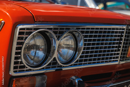 Tight front-quarter view of classic car showing chrome grille with horizontal bars, paired round headlights with reflective lenses, bright red painted hood, polished bumper details and strong contrast