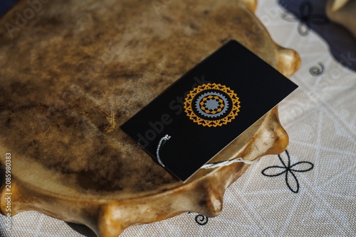 Closeup view of black rectangular gift tag featuring circular geometric gold and gray emblem resting on weathered wooden tray over textured fabric, with warm soft daylight and shallow depth of field