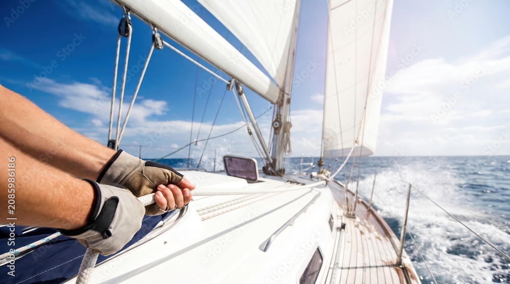 Fototapeta premium A person is pulling ropes on a sailboat while sailing through the ocean. The sky is bright and sunny with some clouds. The water is choppy