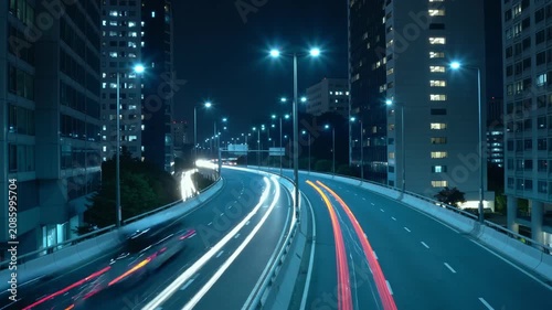 Night highway timelapse shows moving cars and buses between tall city buildings. Bright light trails and curved road create dynamic urban motion scene.