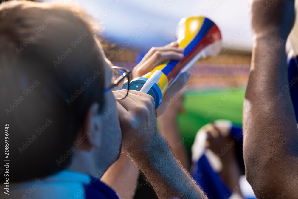 Obraz premium Soccer fans sing with vuvuzelas in stadium tribunes, blowing horns and chanting loudly to motivate their favorite football team.