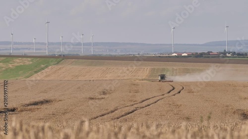 Wide Shot of Combine Harvester Harvesting Wheat Field with Wind Turbines in Background | Handeheld
