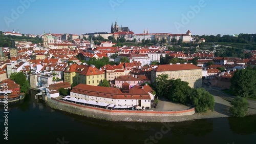 Aerial view of Prague showcasing the vibrant red rooftops, historic buildings, and the majestic St. Vitus Cathedral against a clear blue sky over the Vltava River