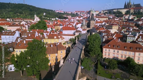 Aerial view of Charles Bridge in Prague, showcasing pedestrians walking along the bridge with historic buildings and lush greenery in the background