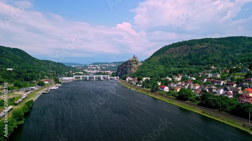 Aerial view of Strekov Castle overlooking the Elbe River, surrounded by lush green hills and residential buildings under a bright blue sky in the Czech Republic