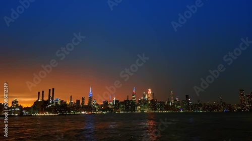 Illuminated Manhattan skyline at night across the East River. City skyscrapers lit up under dark blue twilight sky with water reflection.