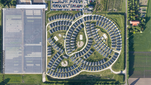 Aerial view of a modern residential neighborhood with a unique curved layout, adjacent to a large glass greenhouse complex and surrounding agricultural fields in Venlo, Limburg, Netherlands.