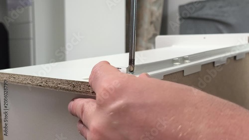 Hand assembling furniture by tightening a screw with a screwdriver on a white particleboard panel, securing a drawer rail and roller component