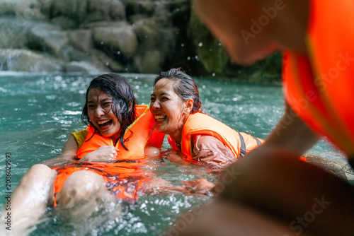 Young Asian people friends wearing life jackets and playing water together at waterfall in tropical forest, enjoying adventure outdoor active travel lifestyle nature therapy on summer holiday vacation