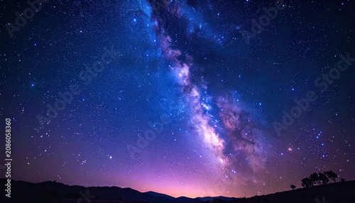 The vast and colorful Milky Way galaxy stretches across the deep blue and purple night sky, illuminating a distant silhouette of rolling hills and scattered trees.