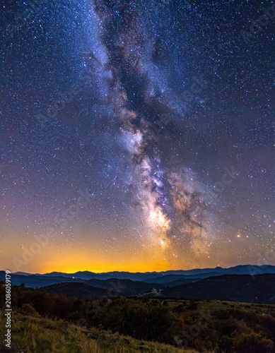 The Milky Way galaxy arcs across a starry night sky over silhouetted hills, with a warm orange glow on the horizon.