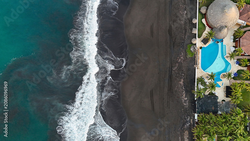 Fotografie Top down drone view of a tropical beachfront resort beside turquoise ocean waves and a black sand beach, with copy space and striking coastal patterns
