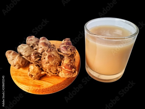 Authentic Indonesian aromatic ginger herbal drink served in a crystal clear glass with fresh aromatic ginger rhizomes placed on a round wooden board, isolated on black background