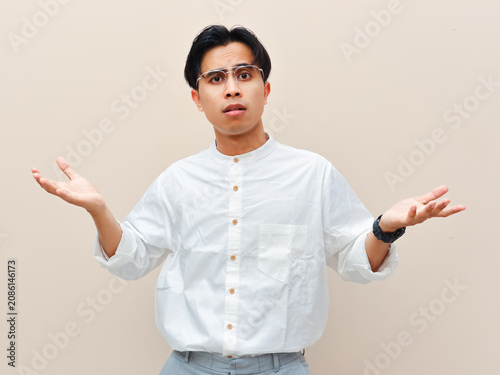 Photography Confused young Asian man shrugging shoulders with open palms gesture against plain background