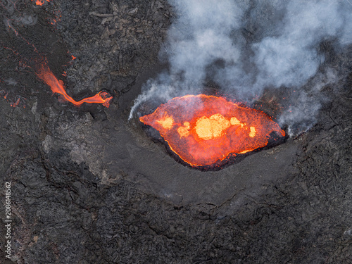 Aerial view of a volcanic eruption with glowing molten lava flowing from a crater surrounded by dark solidified volcanic rock and rising smoke in Iceland.