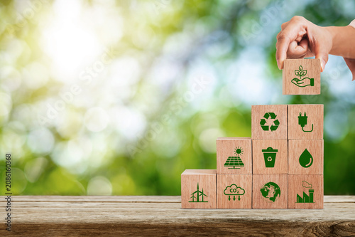A hand placing a wooden block on a pyramid of cubes featuring environmental symbols for carbon reduction, recycling and renewable energy in a natural green setting.