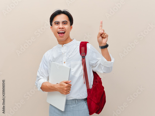 Excited Asian student with backpack holding laptop and pointing finger up.