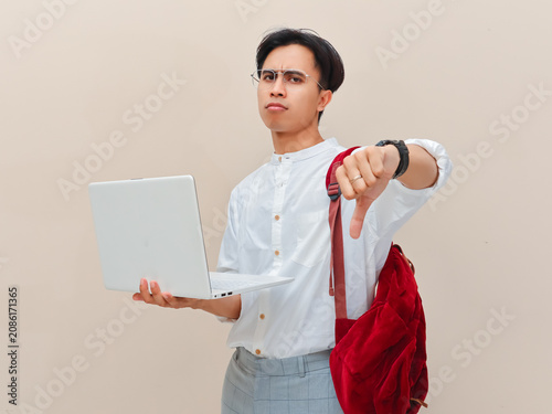 Disappointed Asian student holding open laptop and showing thumbs down sign.