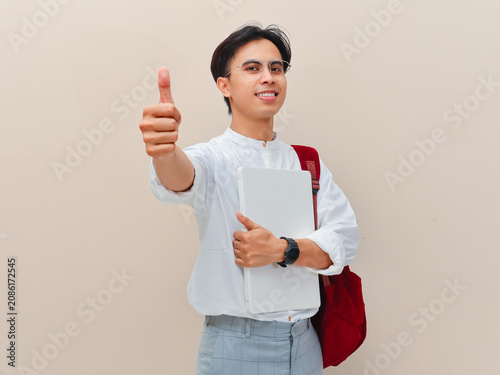 Bild auf Leinwand Cheerful young Asian student or university freshman holding a closed white laptop and gesturing a thumbs up of approval while carrying a red backpack