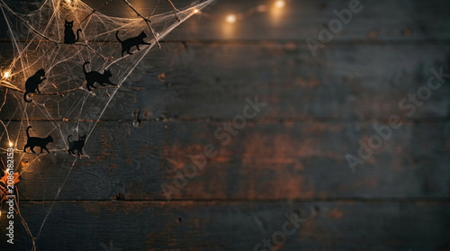 Halloween background with black cat silhouettes on spider webs and branches against a dark horizontal wooden plank wall with glowing yellow fairy lights and empty copy space.