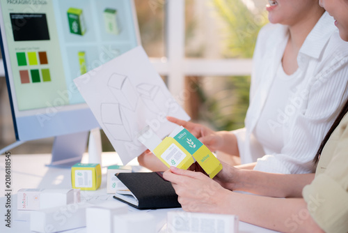 Картината върху платно Two asian woman graphic designers analyzing product package prototype, reviewing paper sketch at home office, comparing physical box mockup with computer screen display with artificial intelligence