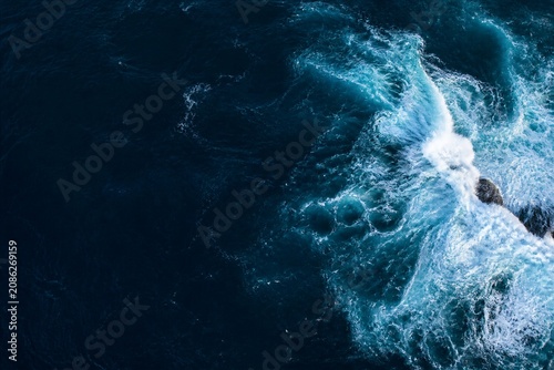 Deep ocean waves crash against rocky shore. Foam explodes in bright white against dark blue. Rocks emerge from churning, powerful water. Aerial view captures nature's raw energy