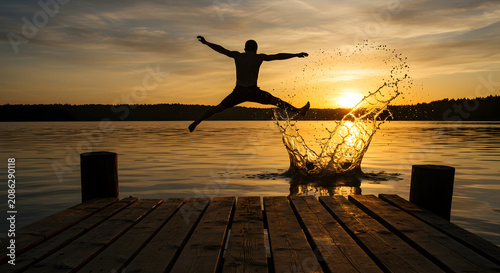 Person Jumping into Water from a Wooden Pier at Sunset with Splashes and Reflection in a Serene Lake Landscape