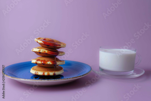 Freshly cooked pancakes stacked on a blue plate drizzled with syrup, accompanied by a glass of milk, on a purple seamless background