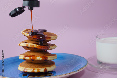 Stacked pancakes drizzling brown syrup from a dispenser, next to a glass of milk, on a purple background