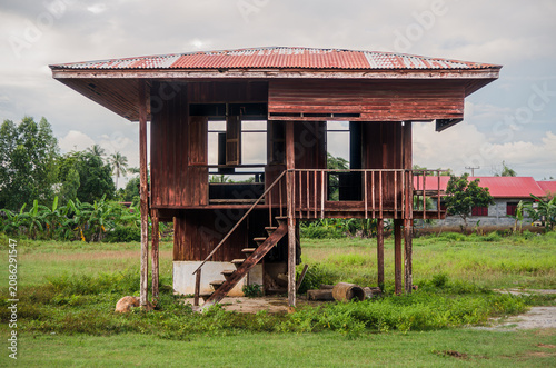 Abandoned Traditional Wooden House on Stilts in Rural Thailand. Old weathered Thai stilt house standing empty and neglected in a grassy field under a cloudy sky in rural Thailand.