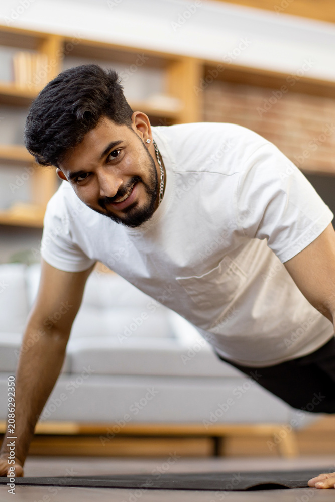 Fototapeta premium A young Indian man with a beard and a white t-shirt performs a push-up exercise on a mat in a modern living room setting