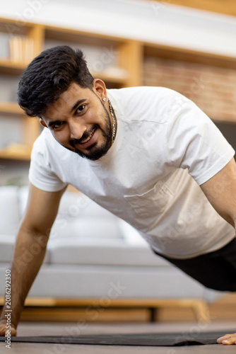 A young Indian man with a beard and a white t-shirt performs a push-up exercise on a mat in a modern living room setting
