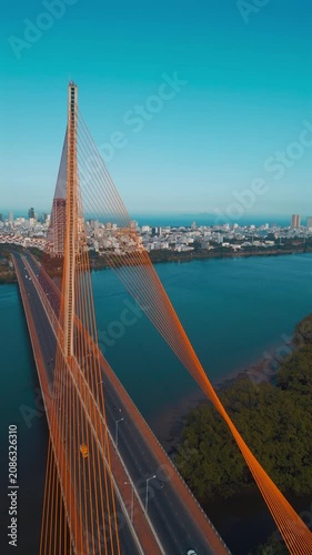 aerial drone view of suspension cable bridge over Han river in Danang, geometric cable-stayed bridge architecture with modern city skyline and roadway, Da Nang Vietnam, 4k vertical video