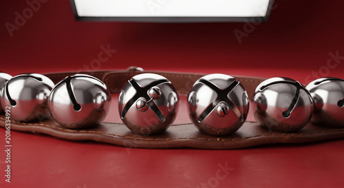 A professional product studio shot of a pristine set of chrome-plated sleigh bells on a new, rich brown leather strap, presented against a seamless, minimalist, festive red background