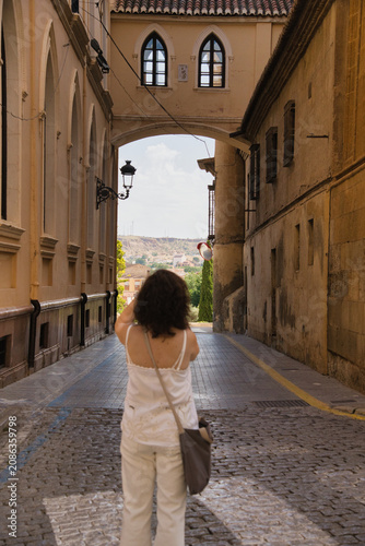 Woman walks through a narrow, historic alleyway. Ancient stone walls frame the path beneath arched openings. Sunlight filters through the arch, casting a warm glow. Her back faces the camera