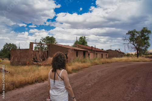 Woman walks alone through abandoned adobe structures. Blue sky with clouds looms overhead. Dusty path leads toward weathered buildings. Dry grass surrounds the silent, sunlit scene