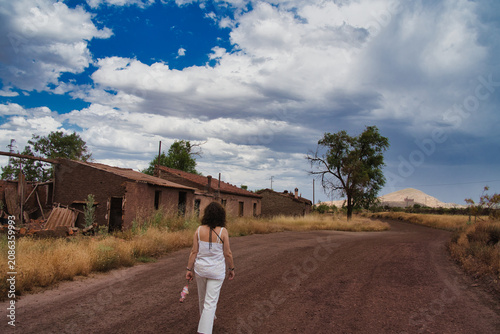 Woman walks down dusty road past weathered adobe homes. Blue sky with clouds looms above arid landscape. Her back is turned as she moves forward toward distant hills