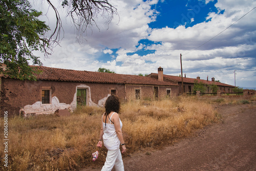 Woman walks past weathered adobe buildings. Dry grass and sparse trees surround the scene. Blue sky with puffy clouds adds contrast. Her gaze suggests quiet contemplation. Ideal for travel, cultural